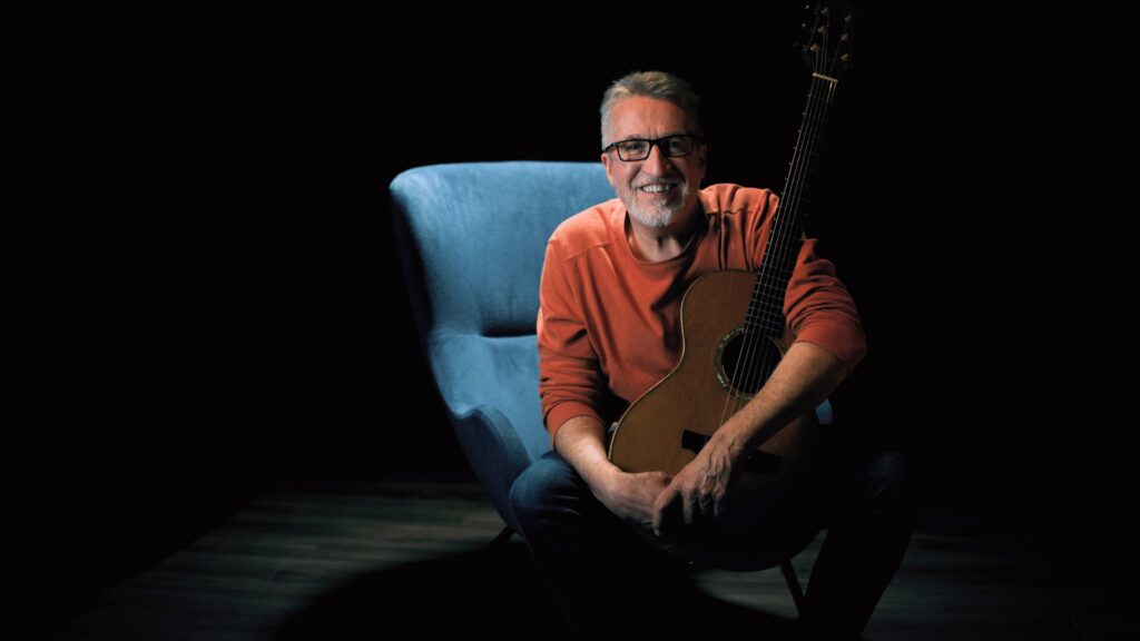 Steve Bell in orange sweater on blue chair with guitar 1