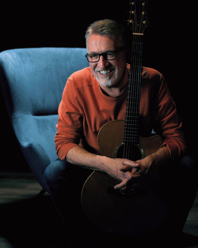 Steve Bell in orange sweater on blue chair with guitar 2