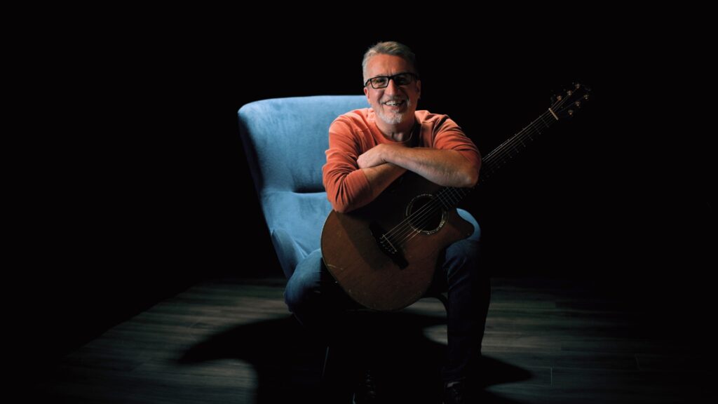 Steve Bell in orange sweater on blue chair with guitar 3