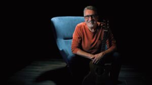 Steve Bell in orange sweater on blue chair with guitar 5