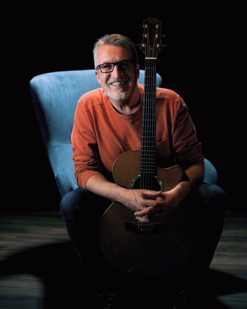 Steve Bell in orange sweater on blue chair with guitar 6