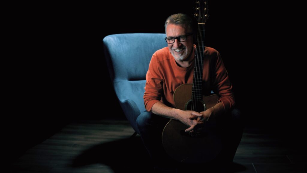 Steve Bell in orange sweater on blue chair with guitar 7