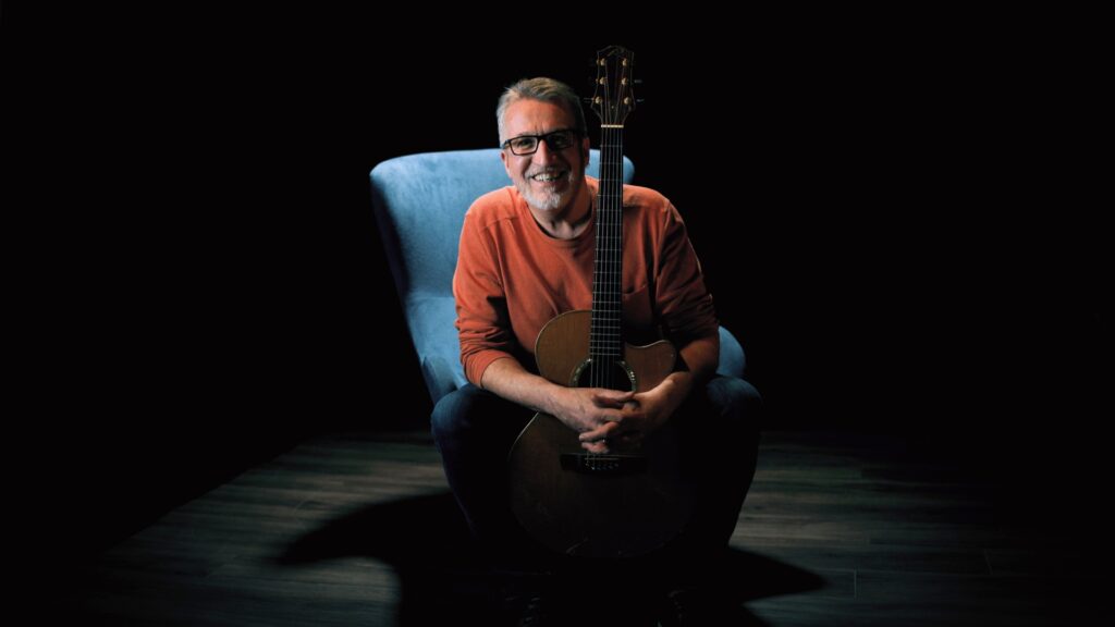 Steve Bell in orange sweater on blue chair with guitar 9